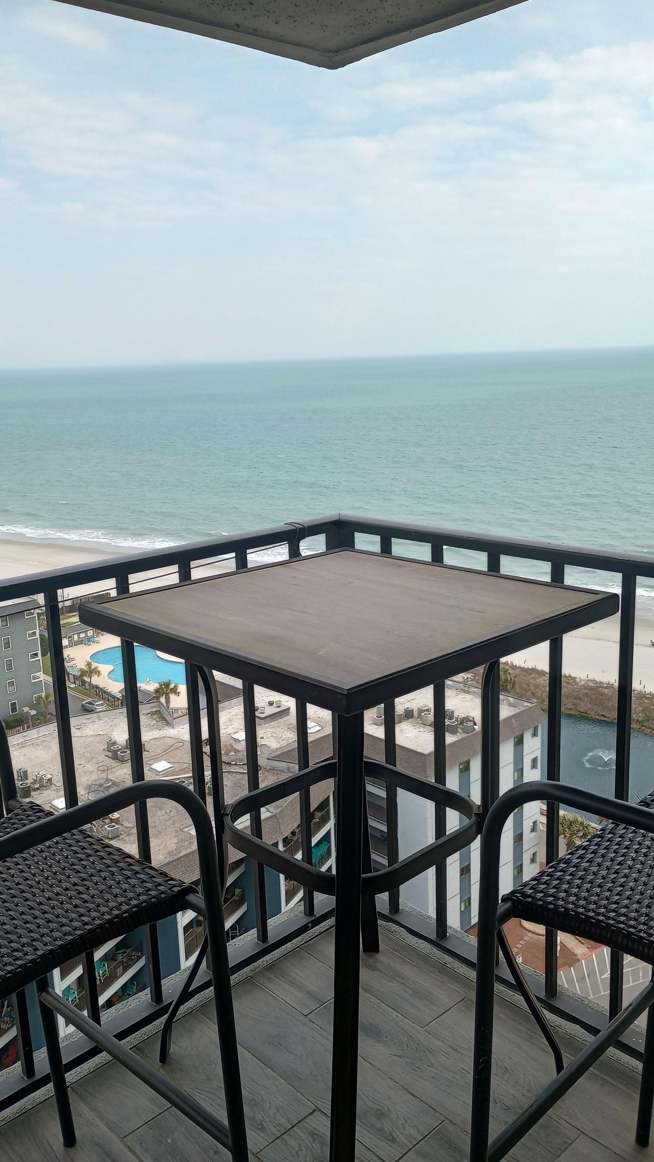 A balcony with a table and two chairs, overlooking a beach and a swimming pool.