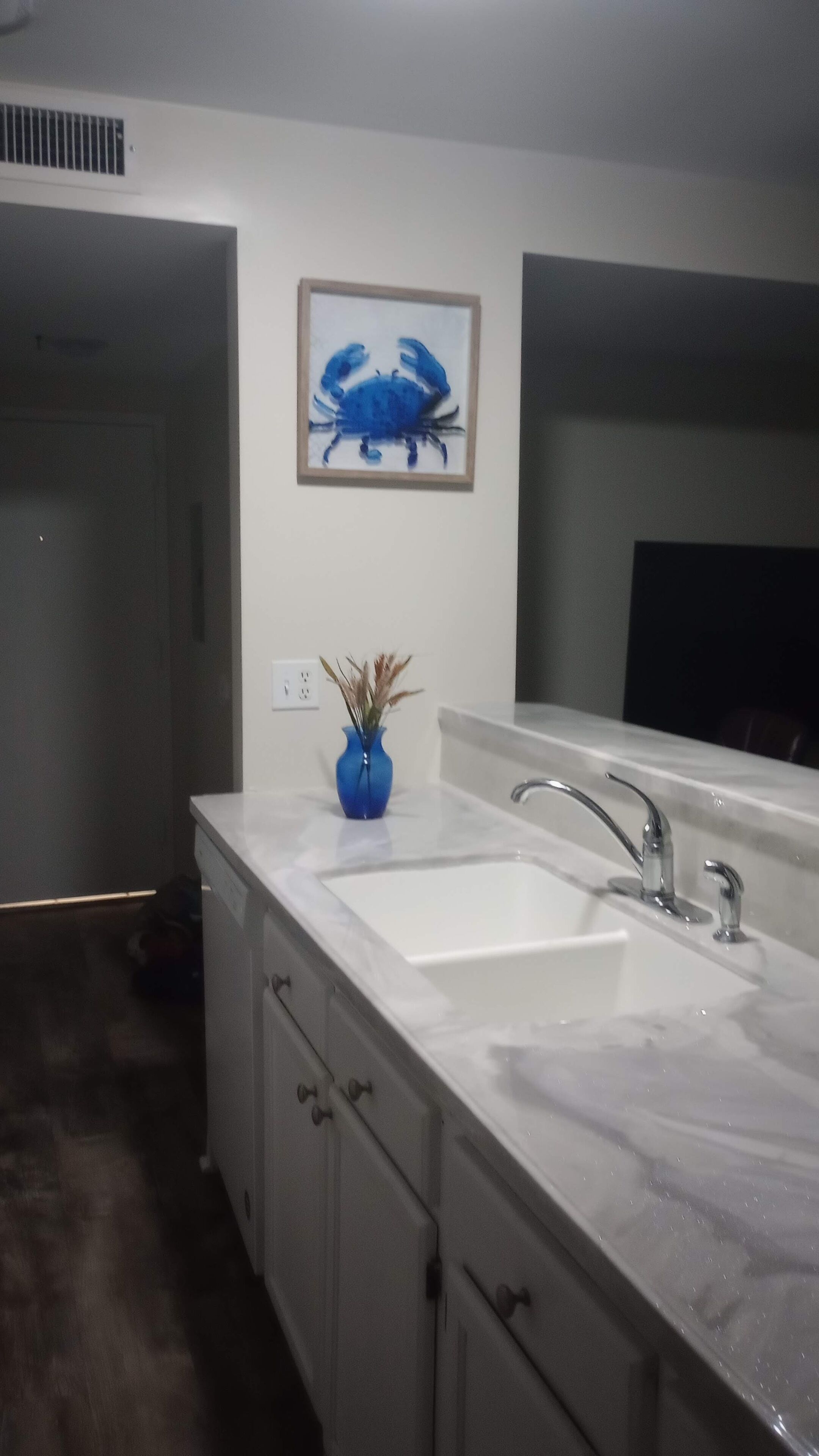 Una cocina moderna con encimera de mármol blanco, dos fregaderos, un jarrón azul con plantas secas y un cuadro con la imagen de un cangrejo en la pared.
