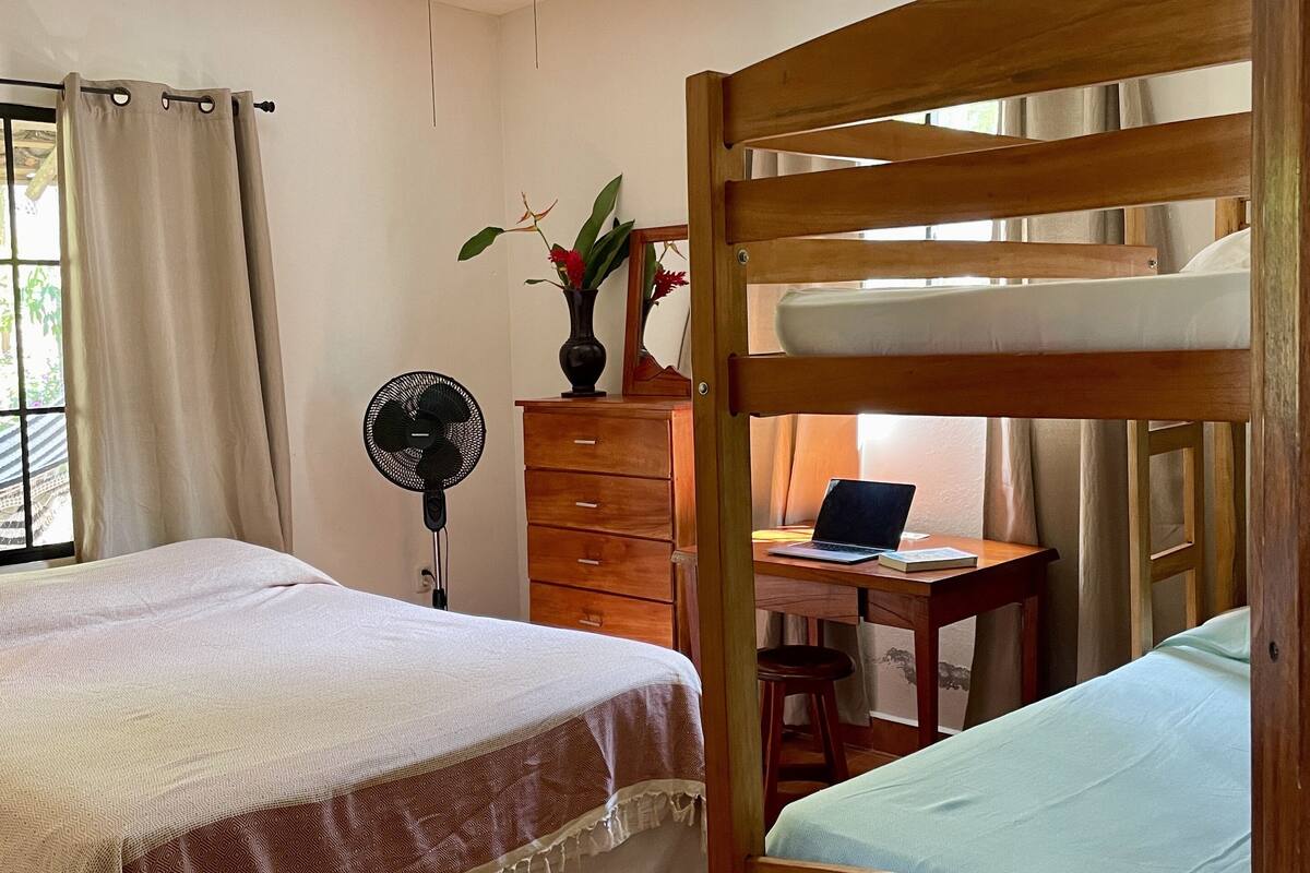 Une chambre avec un lit, un bureau avec un ordinateur portable, un ventilateur et un ventilateur de plafond.