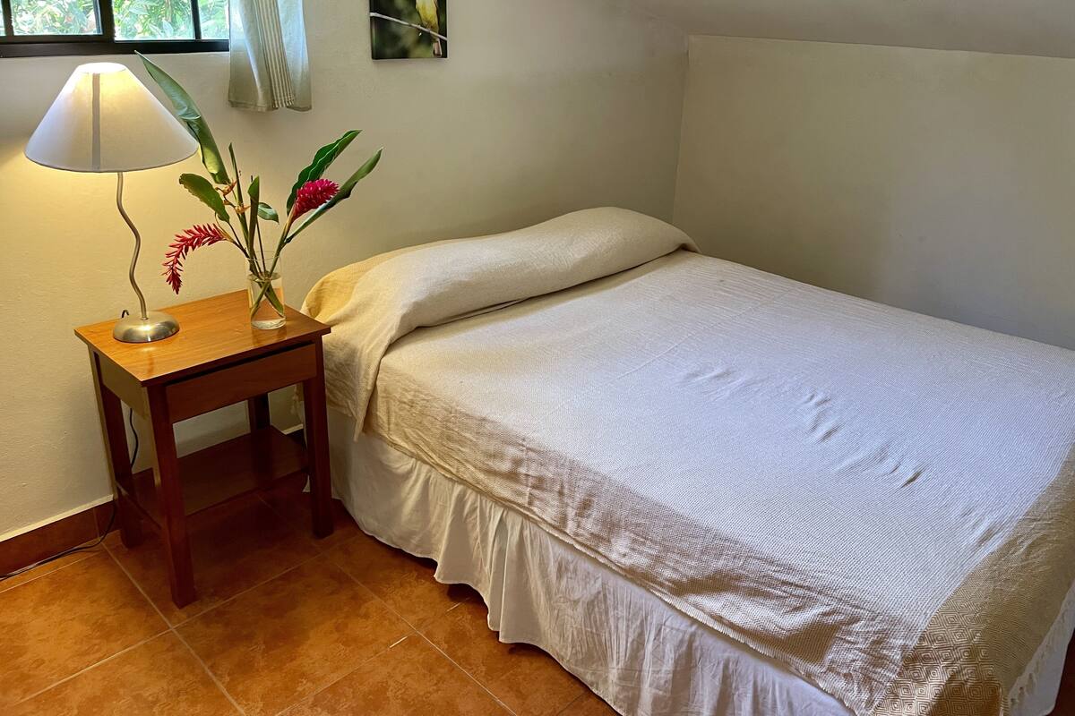 Un lit bien fait, recouvert d’une couvre-lit blanche, dans une chambre où se trouve une table de chevet en bois, sur laquelle reposent une lampe et un vase rempli de fleurs.