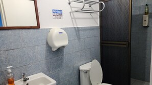 Un baño con lavamanos, inodoro y secamanos. Hay un aviso en la pared y una botella de jabón de manos sobre el lavamanos.