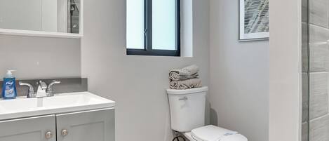 A modern bathroom with a white vanity, a toilet, a window, and a shower area.