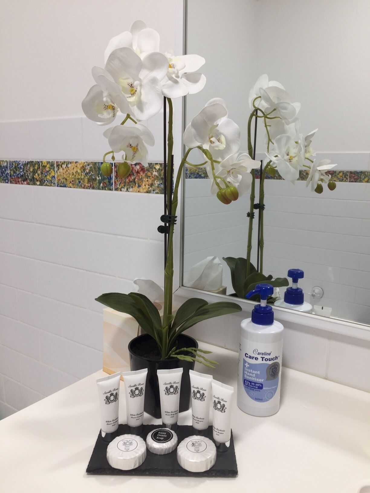 Bagno con piastrelle bianche, uno specchio, orchidee bianche, una pianta in vaso e un espositore di prodotti per la cura della pelle.