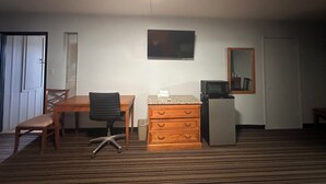 A hotel room with a wooden desk, a black office chair, a small wooden dresser, a television mounted on the wall, and a refrigerator.