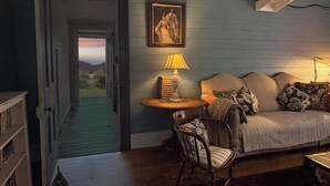 A room with a view through an open door, a framed portrait on the wall, a lamp on a side table, a sofa with cushions, and a chair. - Original 1895 cottage, magnificent uninterrupted views of valley. (Blackheath)