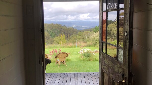 An open wooden door leading to a deck with a wicker chair and a view of a grassy landscape with trees and hills. - Original 1895 cottage, magnificent uninterrupted views of valley. (Blackheath)