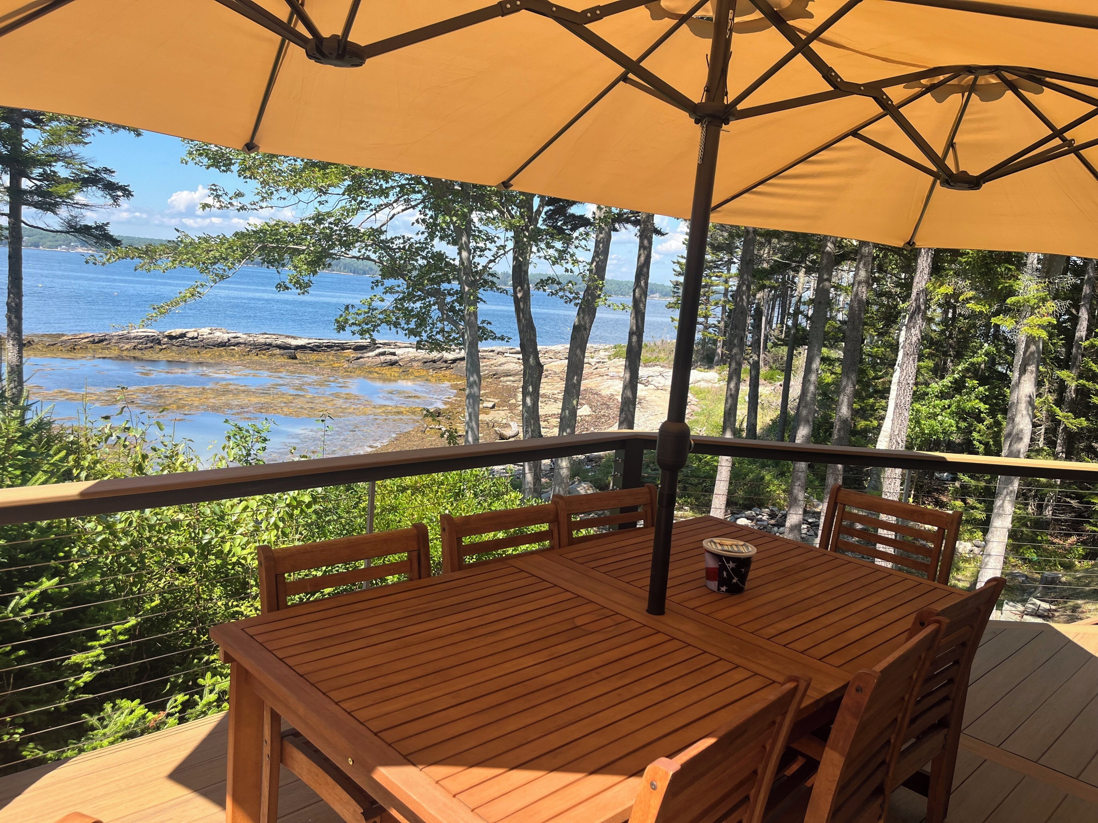 Une terrasse en bois avec une table et des chaises, un parasol, et une vue sur une côte rocheuse et des arbres.