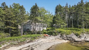 A large, two-story house surrounded by dense forest, with a rocky shoreline and a small body of water.