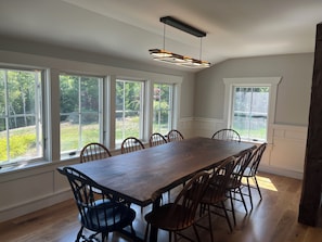 A dining area with a long wooden table and chairs, large windows with a view of greenery, and a modern pendant light fixture.