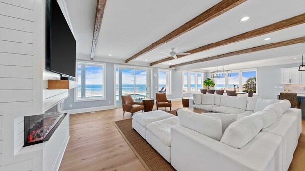 A spacious living room with a large white sectional sofa, wooden beams on the ceiling, and a flat-screen TV mounted on the wall. - Kure Beach Ocean View, Pool & Beach Access (Kure Beach)