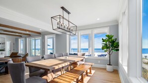 Un vaste espace dédié à la salle à manger, avec une table et des bancs en bois, de grandes fenêtres offrant une vue sur l’océan et une plante en pot.