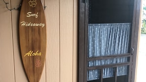 Una tabla de surf de madera con la inscripción 'Surf Hideaway Aloha', una puerta con ventana y un farol pequeño fijado a la pared.