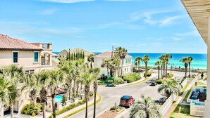 A coastal residential area with houses, palm trees, a road, and a beach in the background.