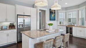A modern kitchen with white cabinetry, a stainless steel refrigerator, a granite countertop, and wicker bar stools. - Rosé Sunsets 6 Seaters Golf Cart and 5 Bikes, Heated Resort Pools, Pickleball Courts (Santa Rosa Beach)