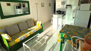 Un espai vital compacte amb un sofà, taula petita de dinar amb cadires, zona de cuina americana i un mirall gran.