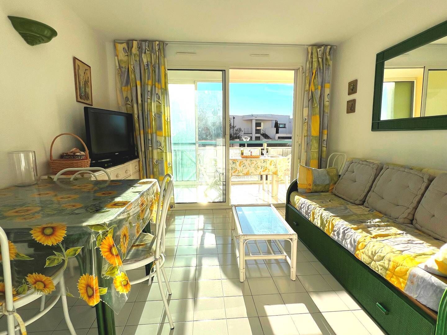 A room with a sofa, a TV, a table with a floral tablecloth, and a balcony with a view.