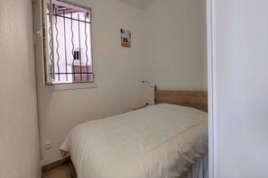 Un dormitorio con una cama individual, una ventana con rejas de metal, un pequeño aire acondicionado de pared y un cabecero de madera.