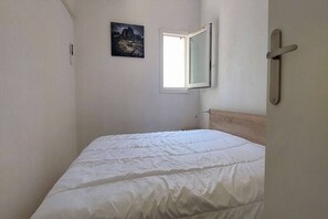 Een kleine, smalle kamer met een enkel bed, een raam en een aan de muur hangend schilderij.