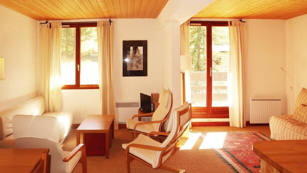 Una habitació acollidora amb un sofà, una taula de fusta, un televisor i una cadira. Hi ha finestres amb cortines, un radiador i un quadre encimbellat a la paret.