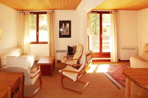 Une pièce confortable avec un canapé, une table en bois, un téléviseur et une chaise. Il y a des fenêtres avec des rideaux, un radiateur et un tableau encadré au mur.