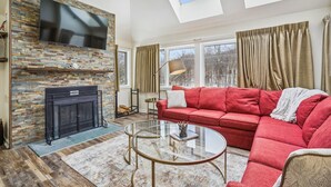A modern living room with a stone fireplace, a red sofa, a glass coffee table, and a TV mounted on a stone wall. - Completely Renovated Three-Bedroom Condo: Your Ideal Retreat A3 (Killington)