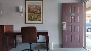 A room with a wooden desk, a chair, a framed landscape painting, a door with a number plate, and a trash bin. - Regency Inn Norco (Norco)