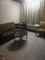 Una sala de estar con un sofá estampado, una mesa redonda de centro, una mesita auxiliar con un teléfono, un equipo de aire acondicionado y una ventana con cortinas.