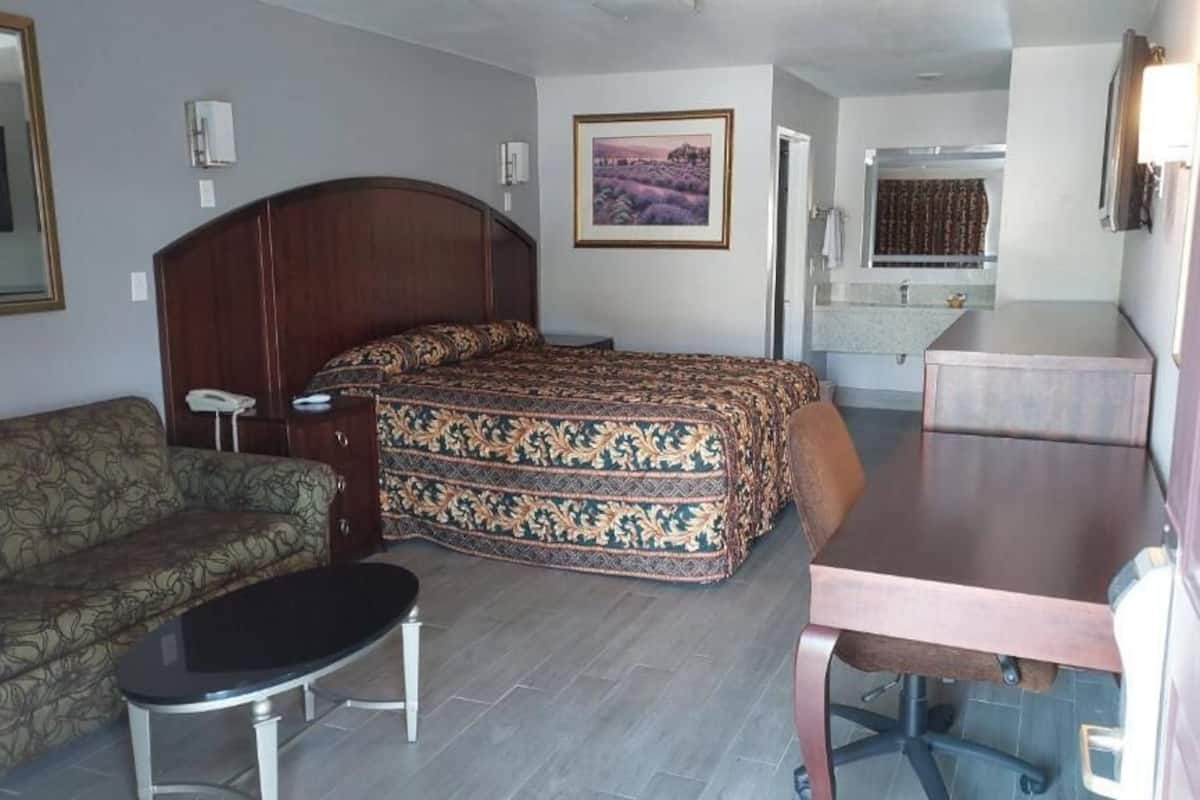 Quarto de hotel compacto com cama, sofá, mesa pequena, escrivaninha e banheiro visível por uma porta aberta.