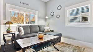A living room with a grey sofa, a coffee table, a lamp, and a view of trees through a window.