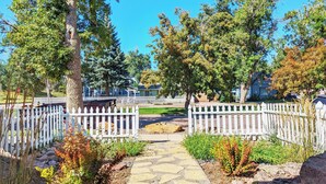 A stone pathway leading to a gated entrance with white picket fences, surrounded by trees and shrubs.