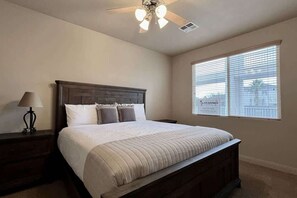 Ein Schlafzimmer mit Bett, Nachttisch, Lampe, Deckenventilator und einem Fenster mit Jalousien.