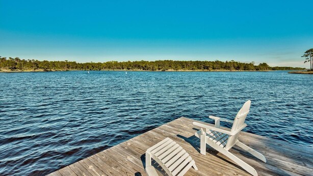 A wooden dock with two white Adirondack chairs facing a calm lake.