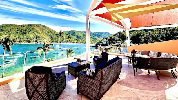 A covered outdoor seating area with wicker furniture, overlooking a pool and a mountainous landscape.