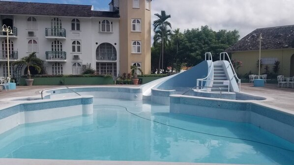 A swimming pool with a slide, surrounded by a building with balconies and a palm tree. - apartment by the beach sleeps 4 (Ocho Rios)