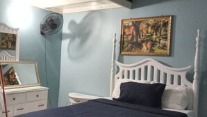 Une chambre à coucher comprenant un lit, une table de chevet, une commode, un miroir, un ventilateur et un tableau accroché au mur.