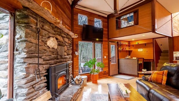 A cozy living room with a stone fireplace, wooden walls, a leather sofa, and a dining area. - Maple Castle – Cozy Riverside Retreat with Hot Tub, Trails & Fireplace (Hancock)