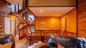 A cozy interior with leather sofas, a wooden dining table, and a staircase leading to an upper level. - Maple Castle – Cozy Riverside Retreat with Hot Tub, Trails & Fireplace (Hancock)