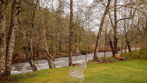 A river flowing through a forest with a swing chair and a bench near the water. - Maple Castle – Cozy Riverside Retreat with Hot Tub, Trails & Fireplace (Hancock)