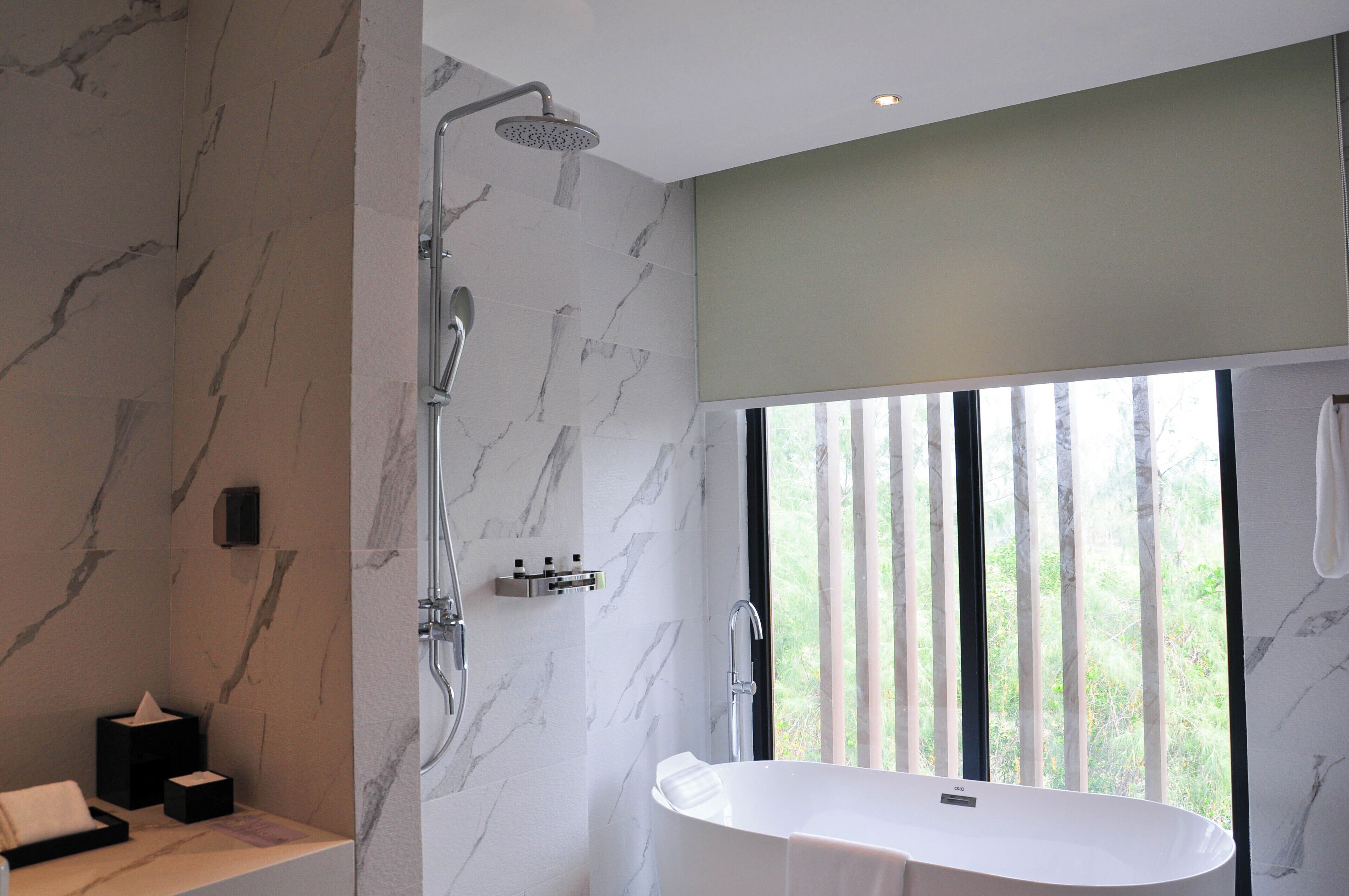 A modern bathroom with a freestanding bathtub, a shower with a rain showerhead, and a large window providing a view of greenery outside.