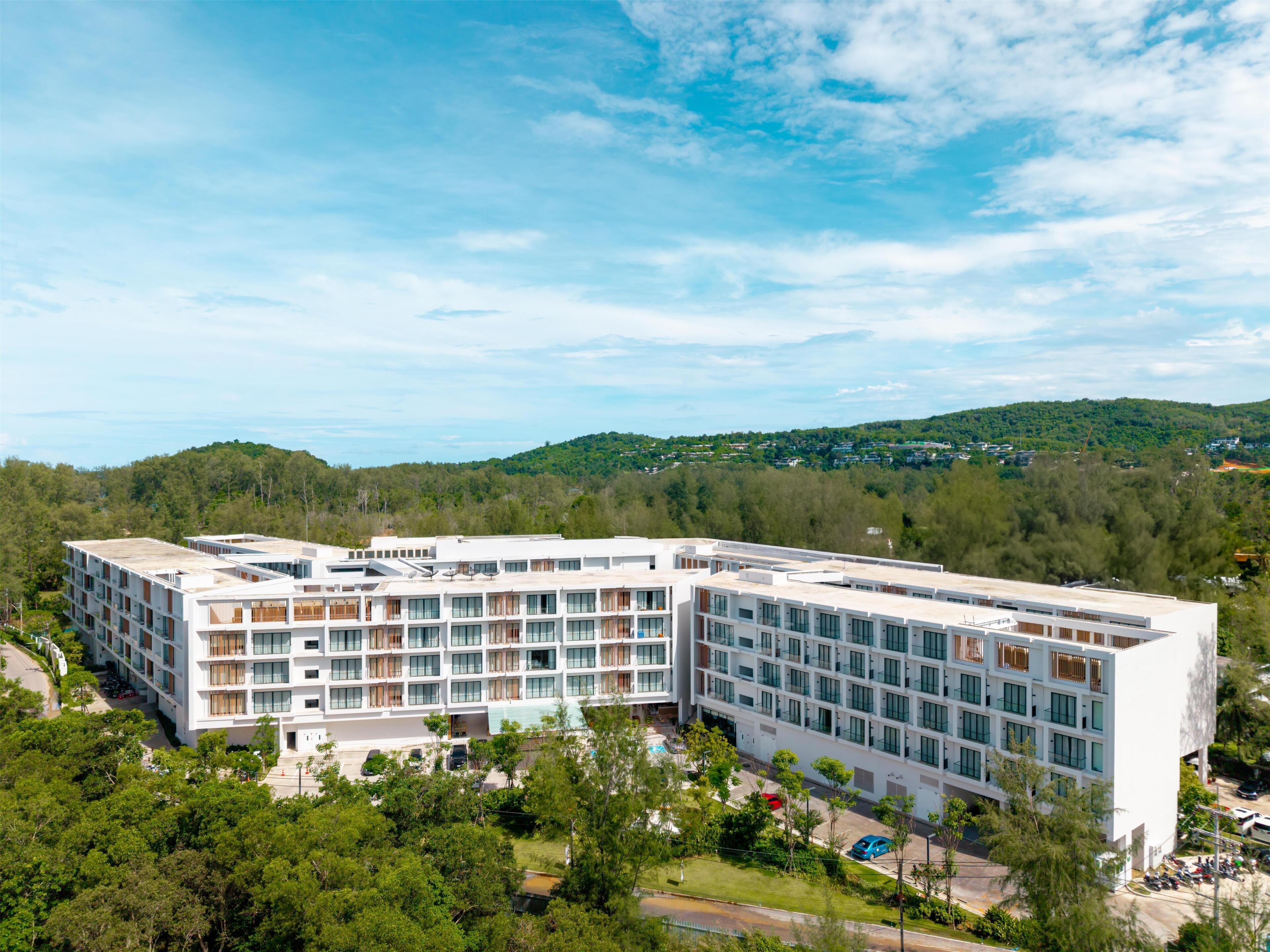 Een modern, meerlaags hotel met een plat dak, omgeven door groen en heuvels op de achtergrond.