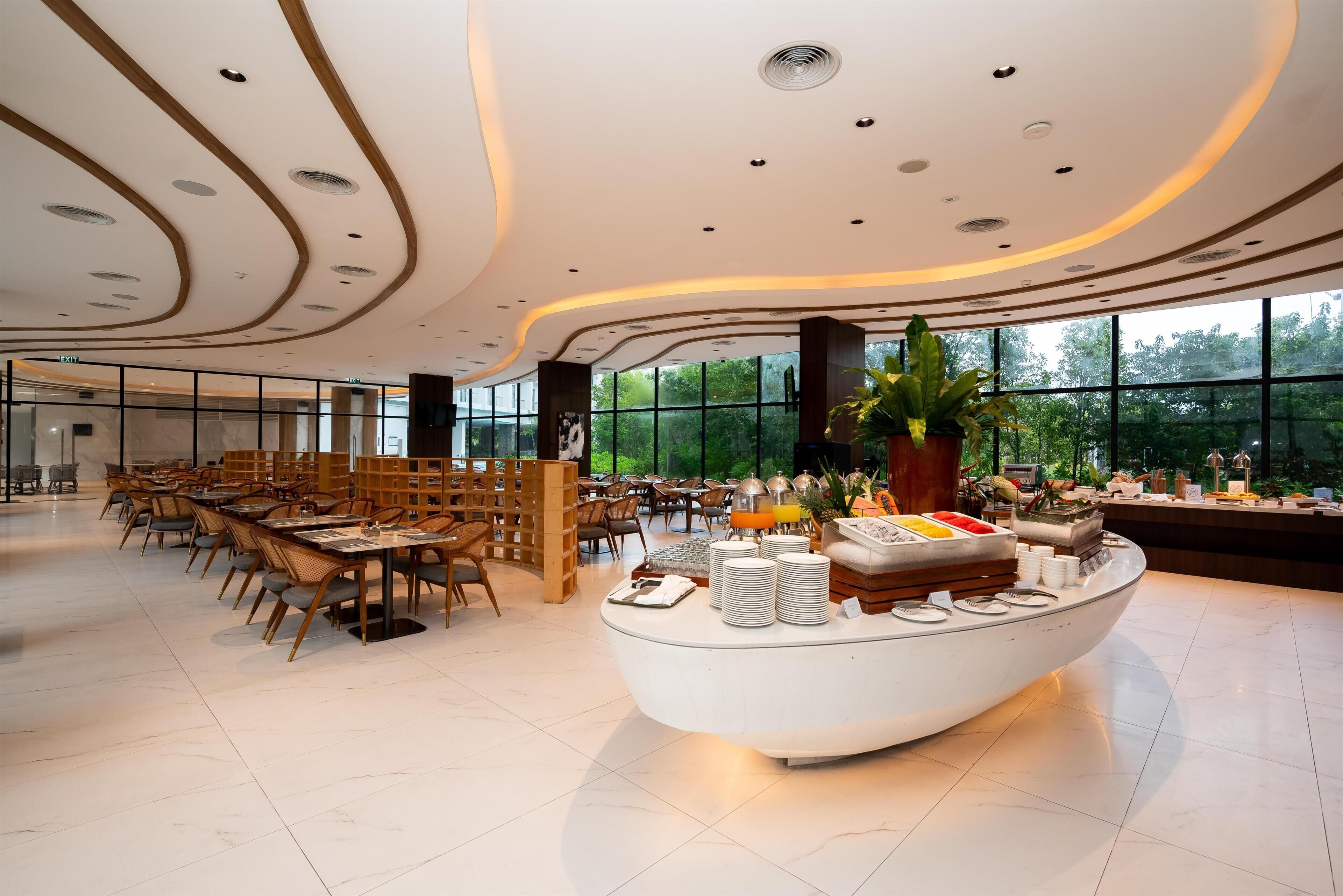 En rymlig hotelllobby med ett centralt placerat buffébord, sittgrupper och stora fönster med utsikt över grönområden.