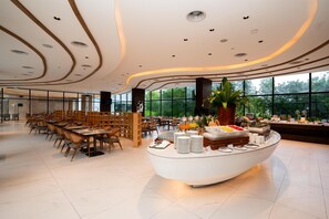 Un amplio vestíbulo de hotel con una zona de buffet central, diferentes disposiciones de asientos y grandes ventanales que ofrecen vistas a zonas verdes.