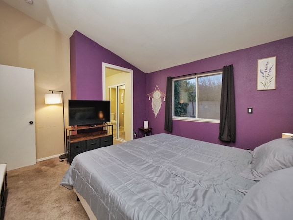A bedroom with a bed, a TV on a stand, a window with curtains, and a door leading to another room.