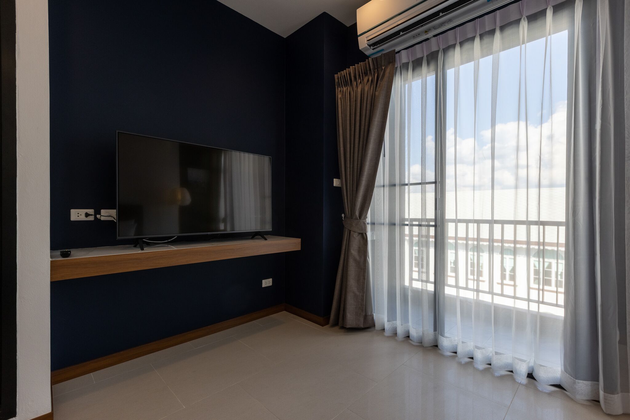 Una habitación con televisión de pantalla plana sobre una repisa de madera, un ventanal con cortinas translúcidas y vistas al cielo y edificios.