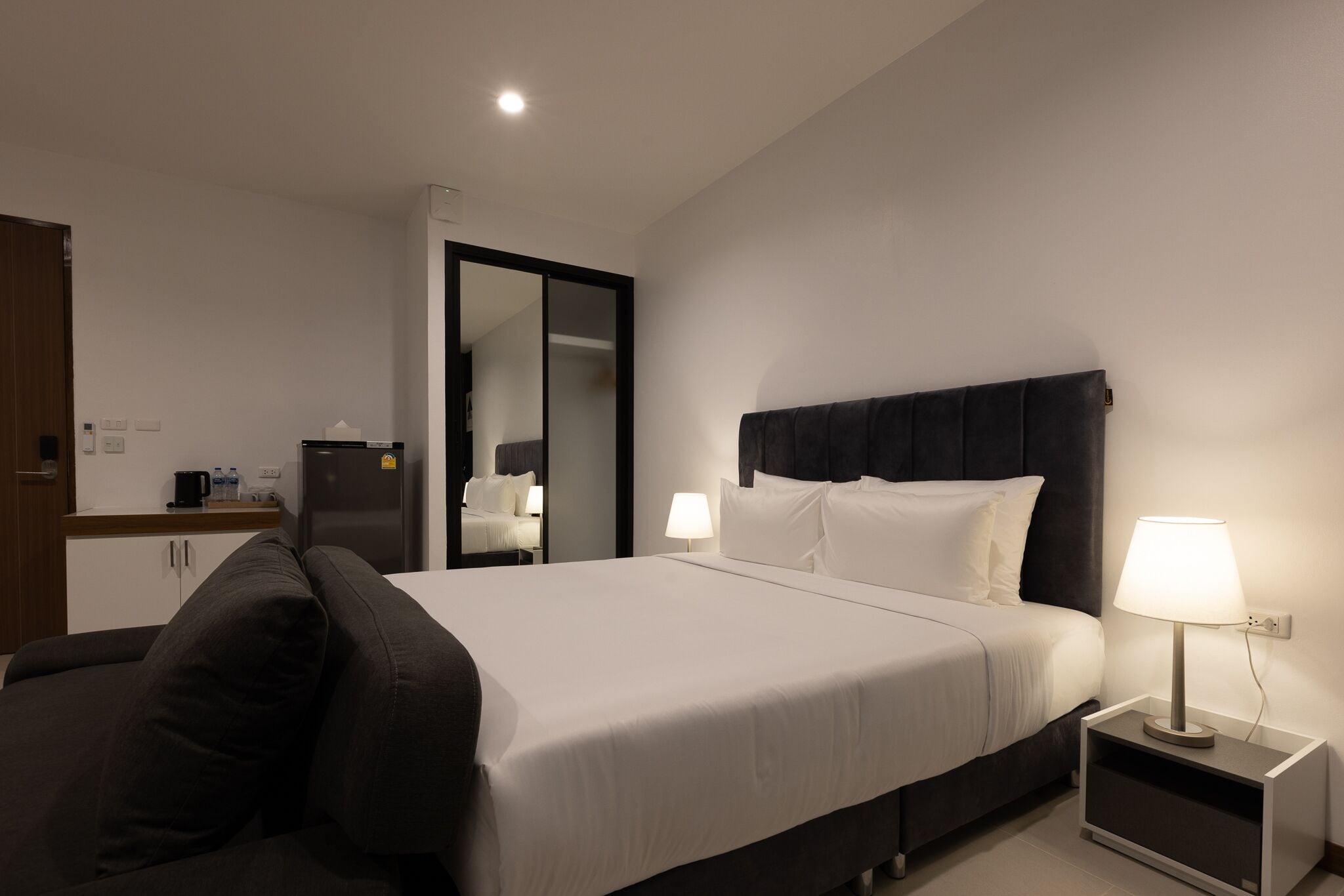 Habitación de hotel con cama, mesita de noche con lámpara, espejo, armario y una pequeña cocina.