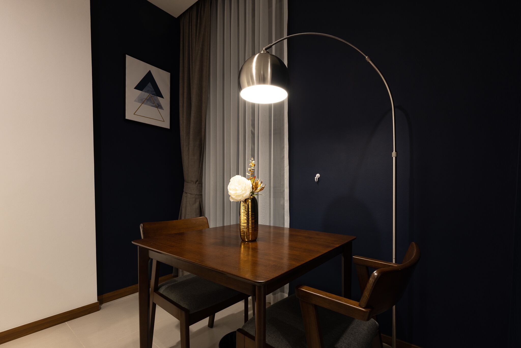 A dining area with a table, chairs, a vase with flowers, a lamp, and a framed picture on the wall.