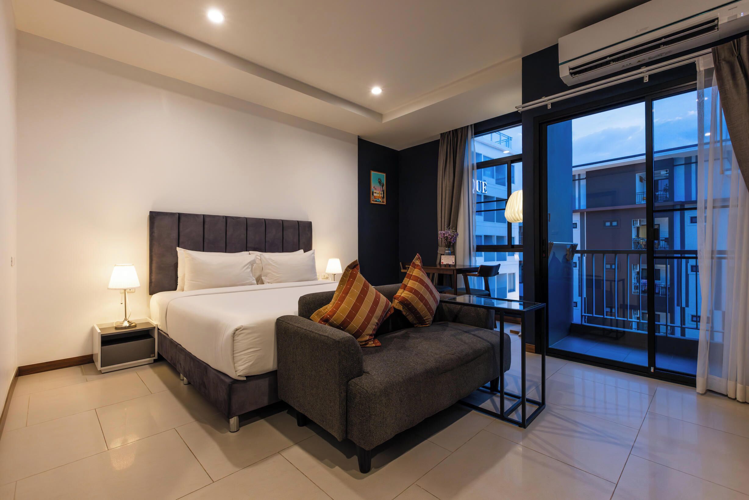 A hotel room with a bed, a sofa, a nightstand, and a large window with a view of buildings.