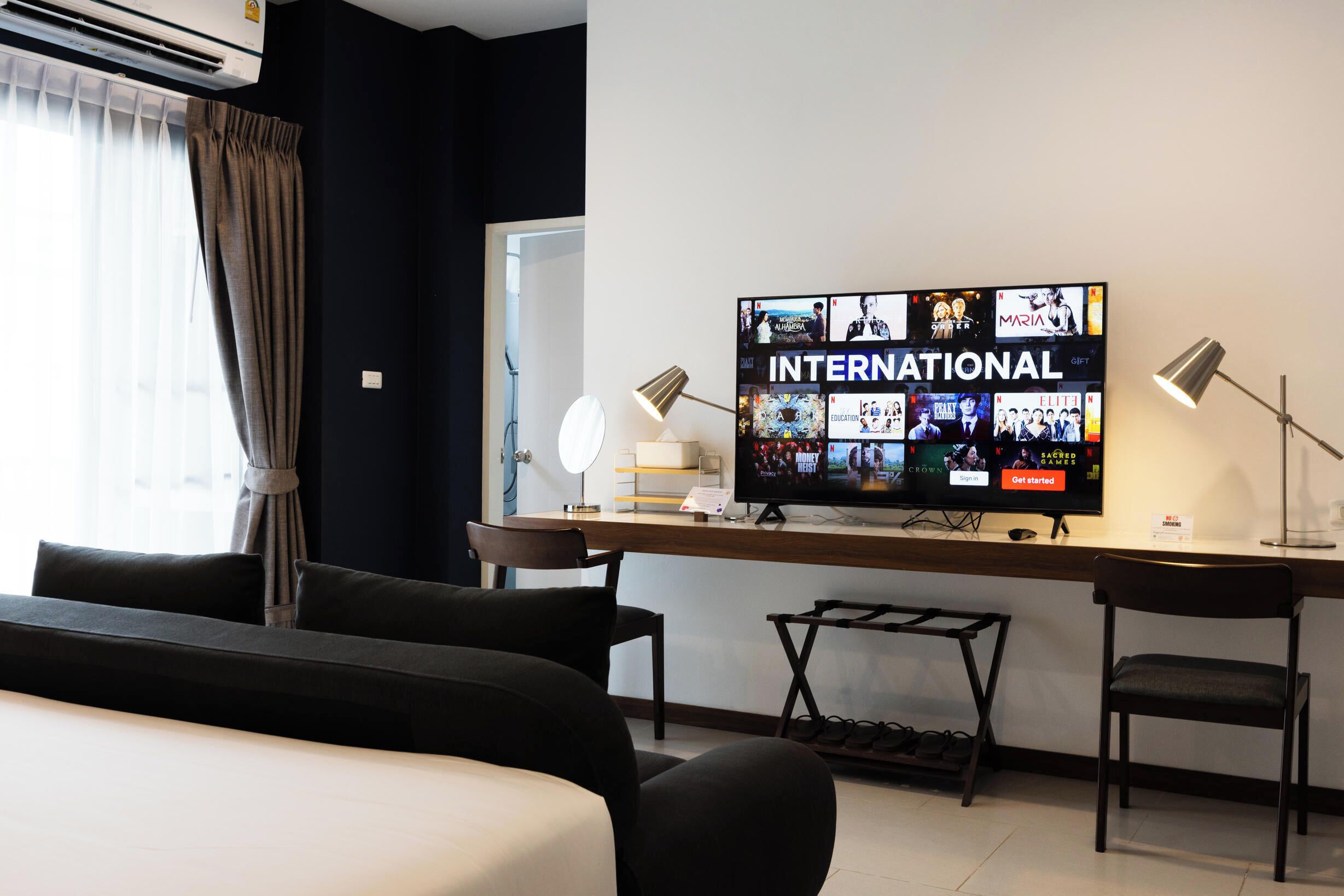 Une chambre d’hôtel dotée d’un grand téléviseur à écran plat diffusant diverses chaînes, d’un bureau avec une lampe, d’une chaise et d’un lit agrémenté d’oreillers de couleur sombre.