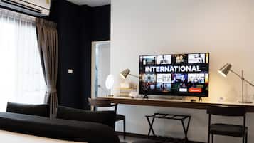 A hotel room with a large flat-screen TV displaying various channels, a desk with a lamp, a chair, and a bed with dark-colored pillows.