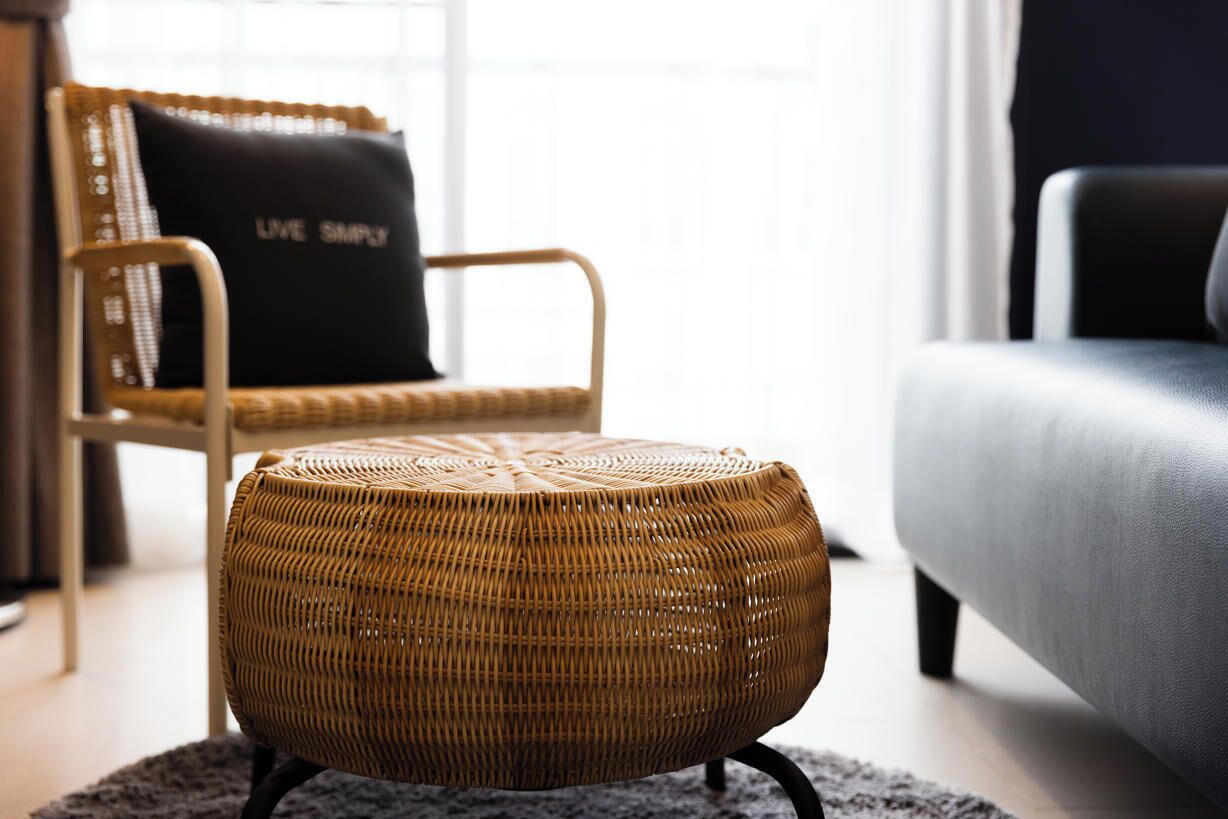 A wicker ottoman on a rug, a wicker chair with a black cushion, and a grey sofa in a living room.